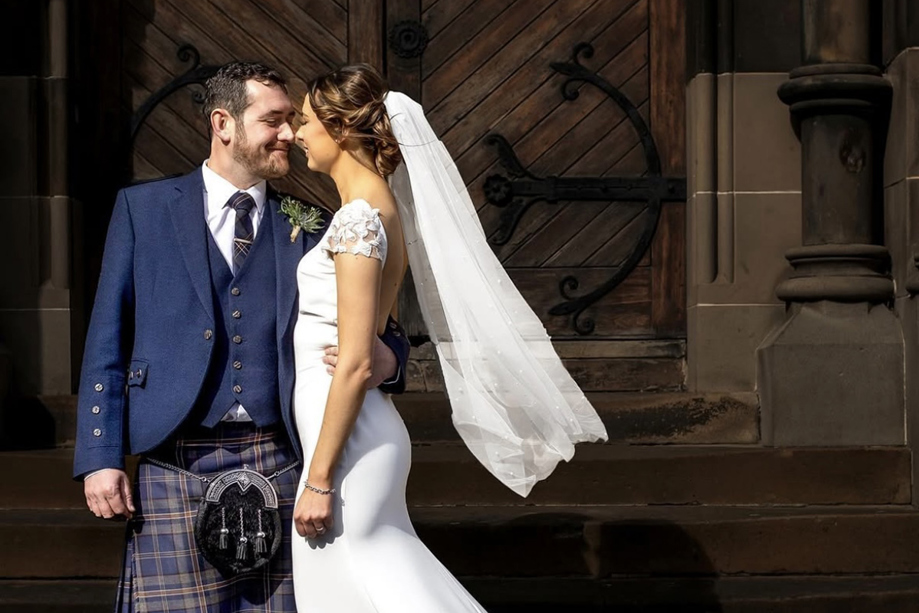 Bride with veil and groom wearing a kilt standing outside venue in an embrace while smiling at each other